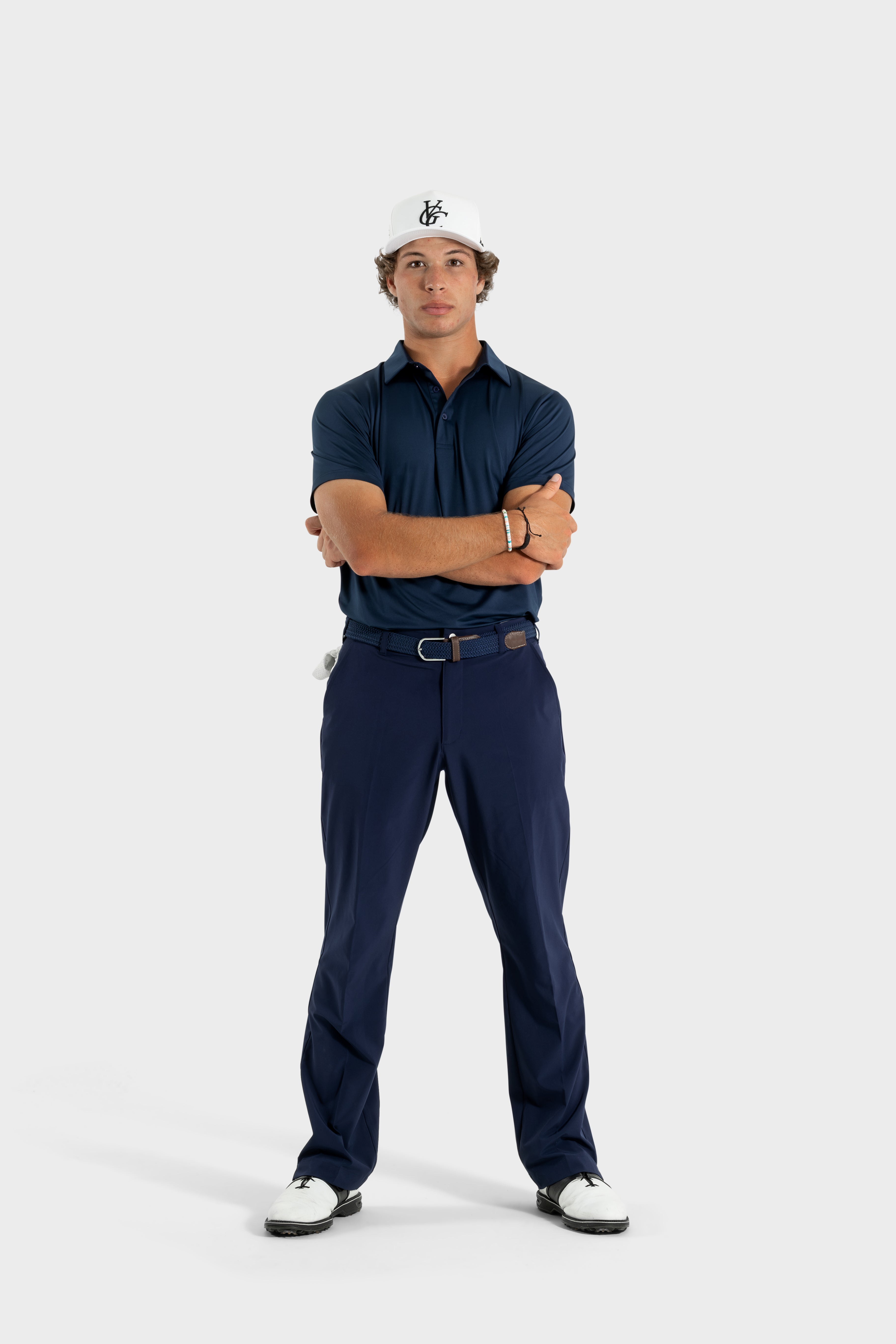 Man wearing The Abyss Golf Polo in navy blue with matching pants and white cap, posing with arms crossed against a white background