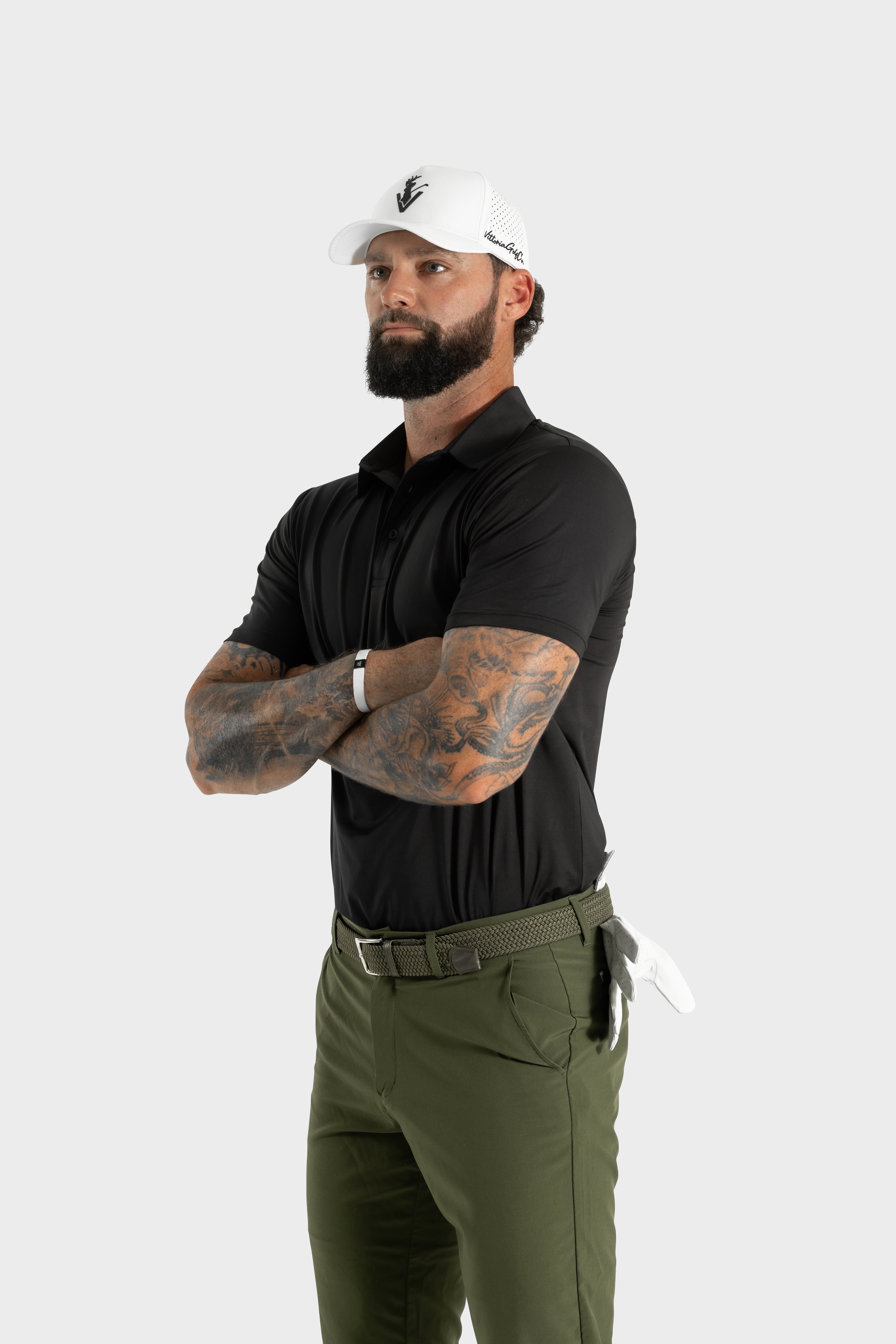Male model wearing The Nothing polo black golf shirt with olive green pants and white cap standing with arms crossed