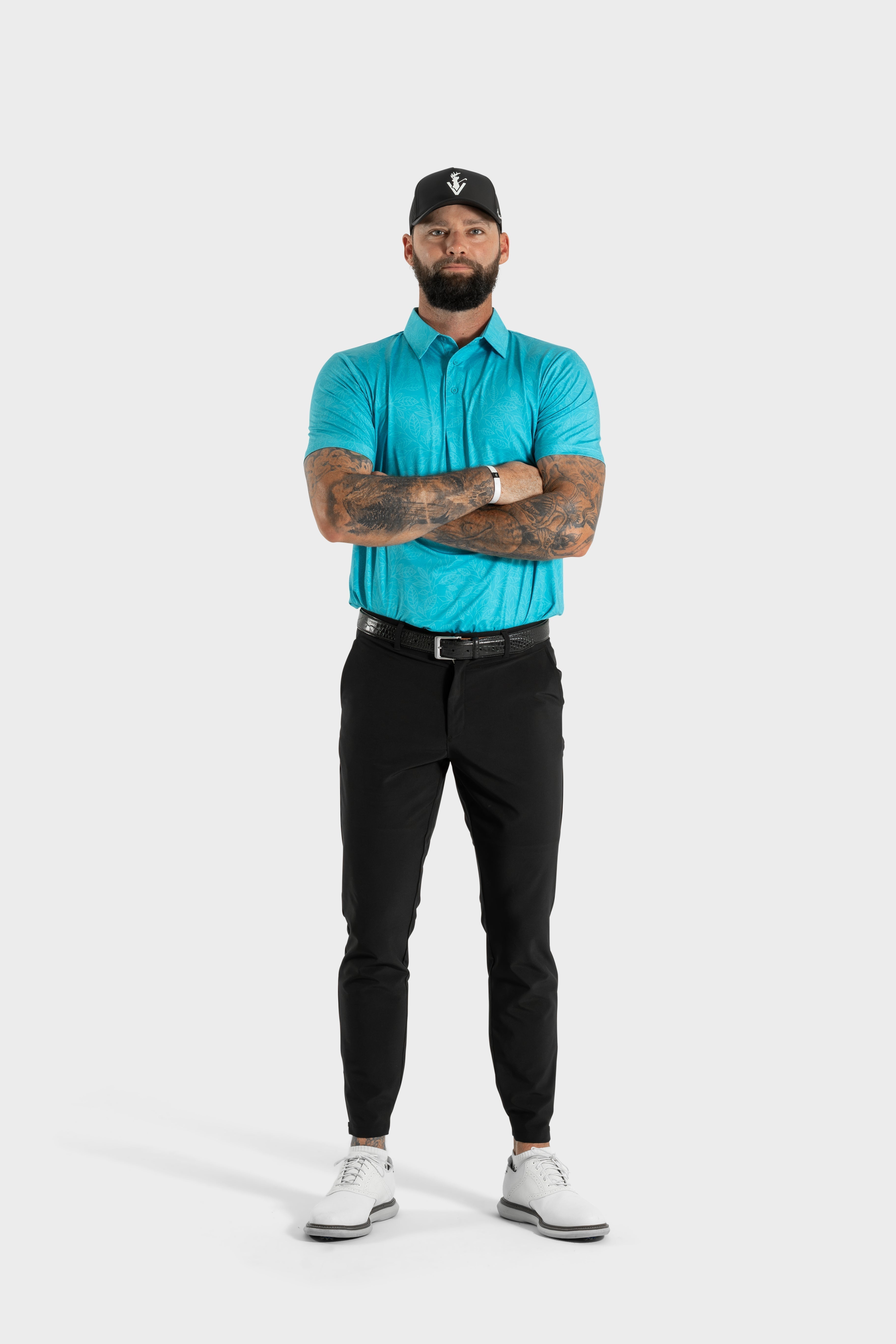 Man wearing turquoise Silent Bloom polo with black pants and white shoes standing with arms crossed