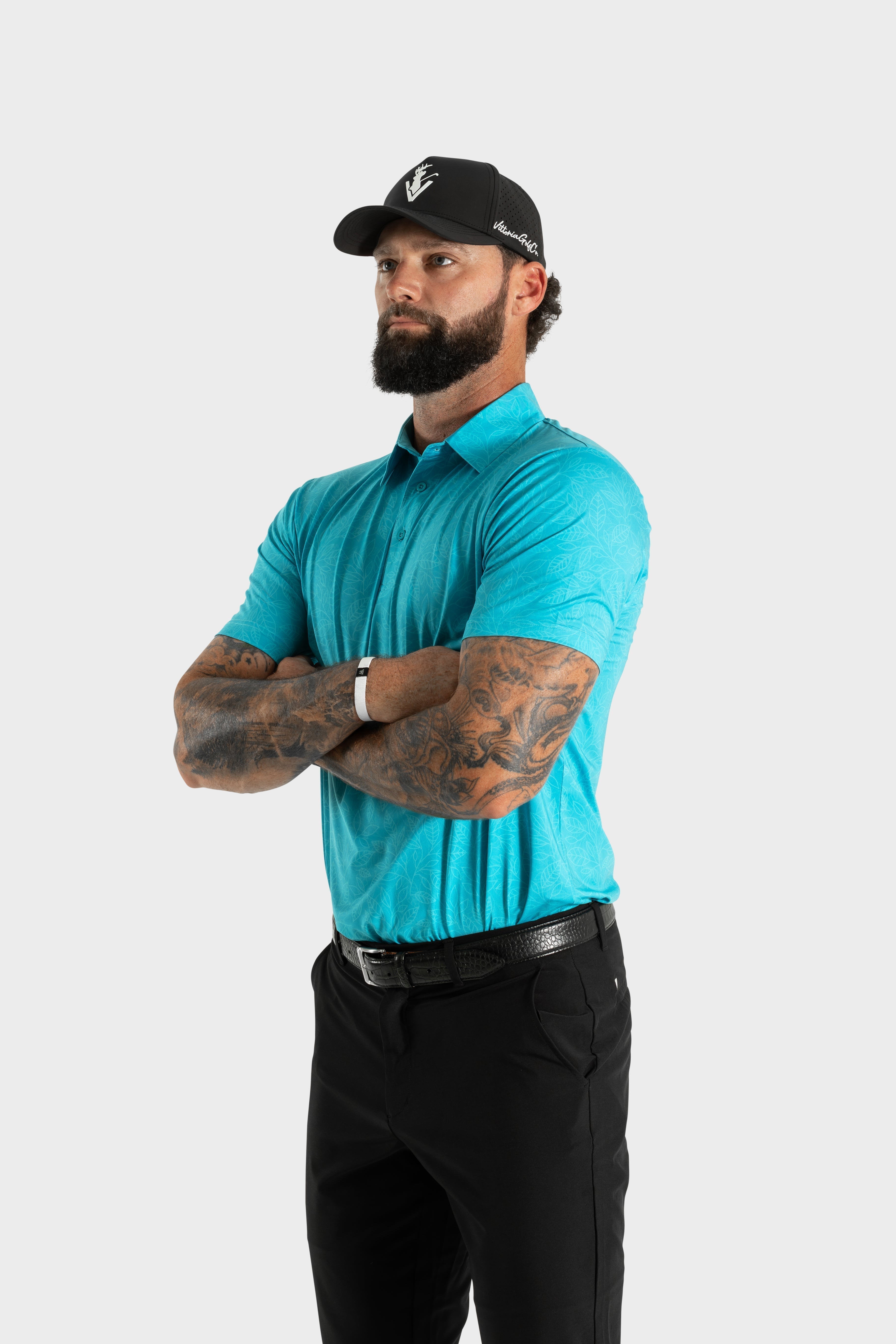 Man wearing turquoise Silent Bloom polo shirt with black pants and cap standing with arms crossed