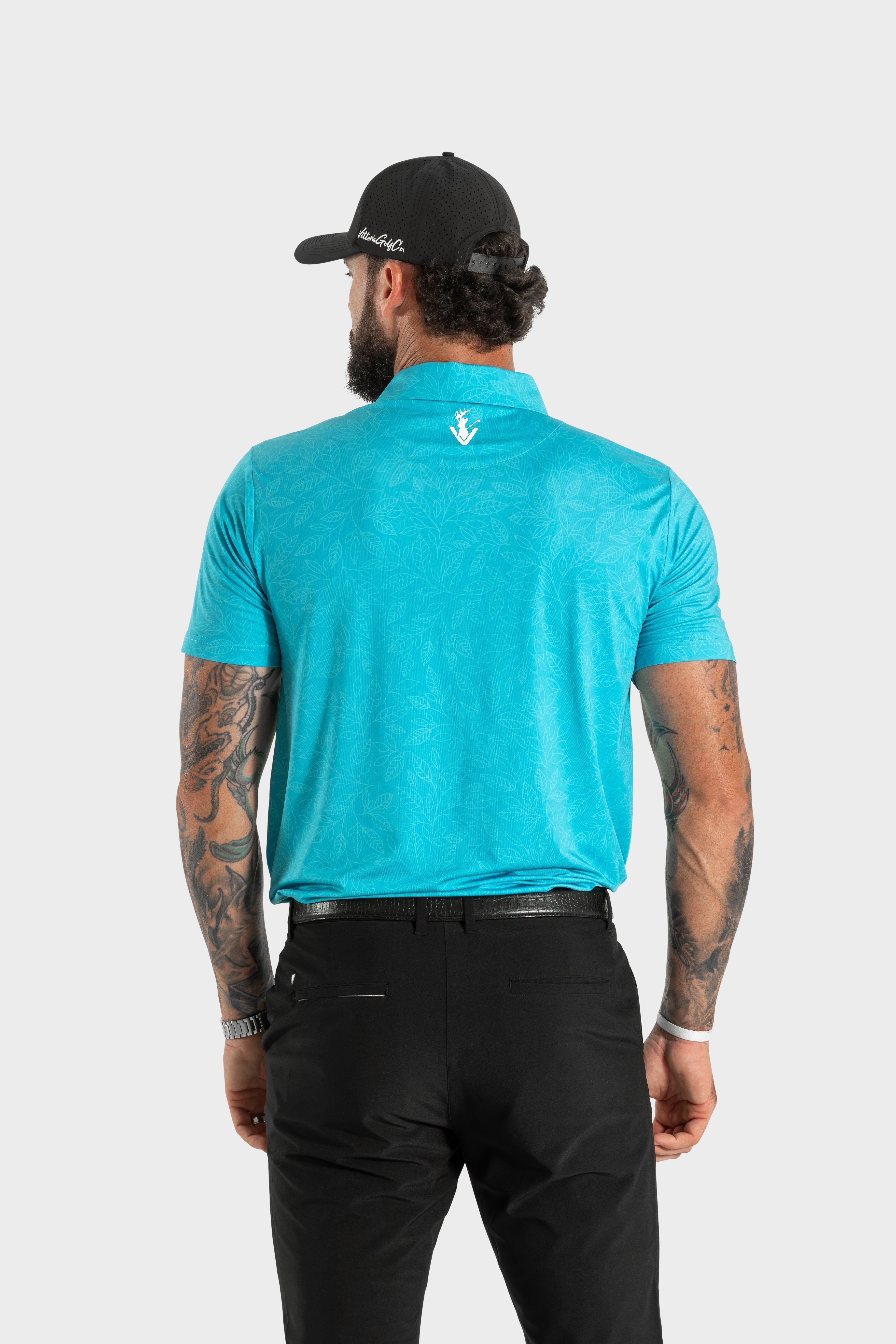 Man wearing turquoise Silent Bloom polo with black pants and cap, back view