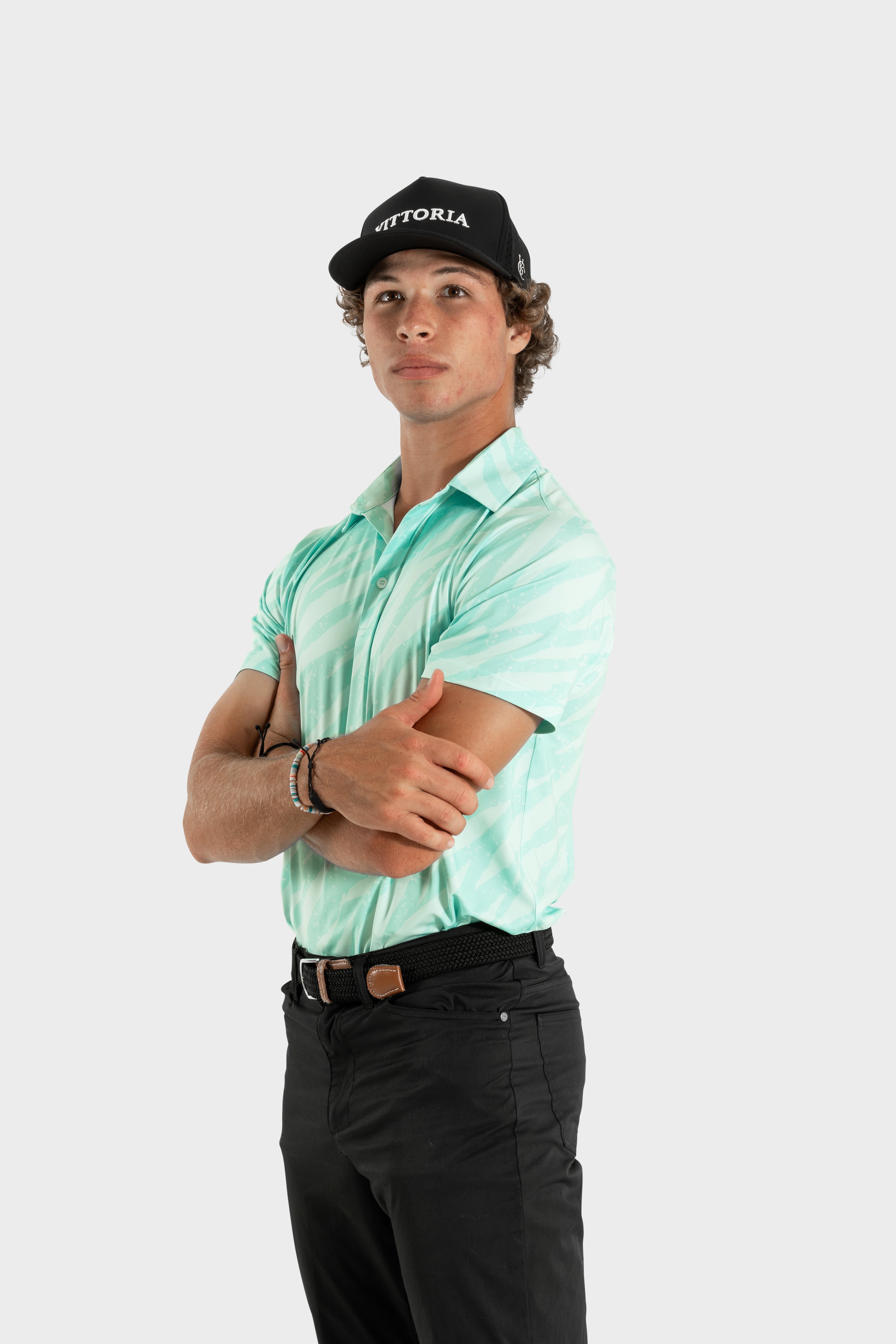 Young man wearing The TigerMint mint-green polo shirt with black pants and cap standing with arms crossed
