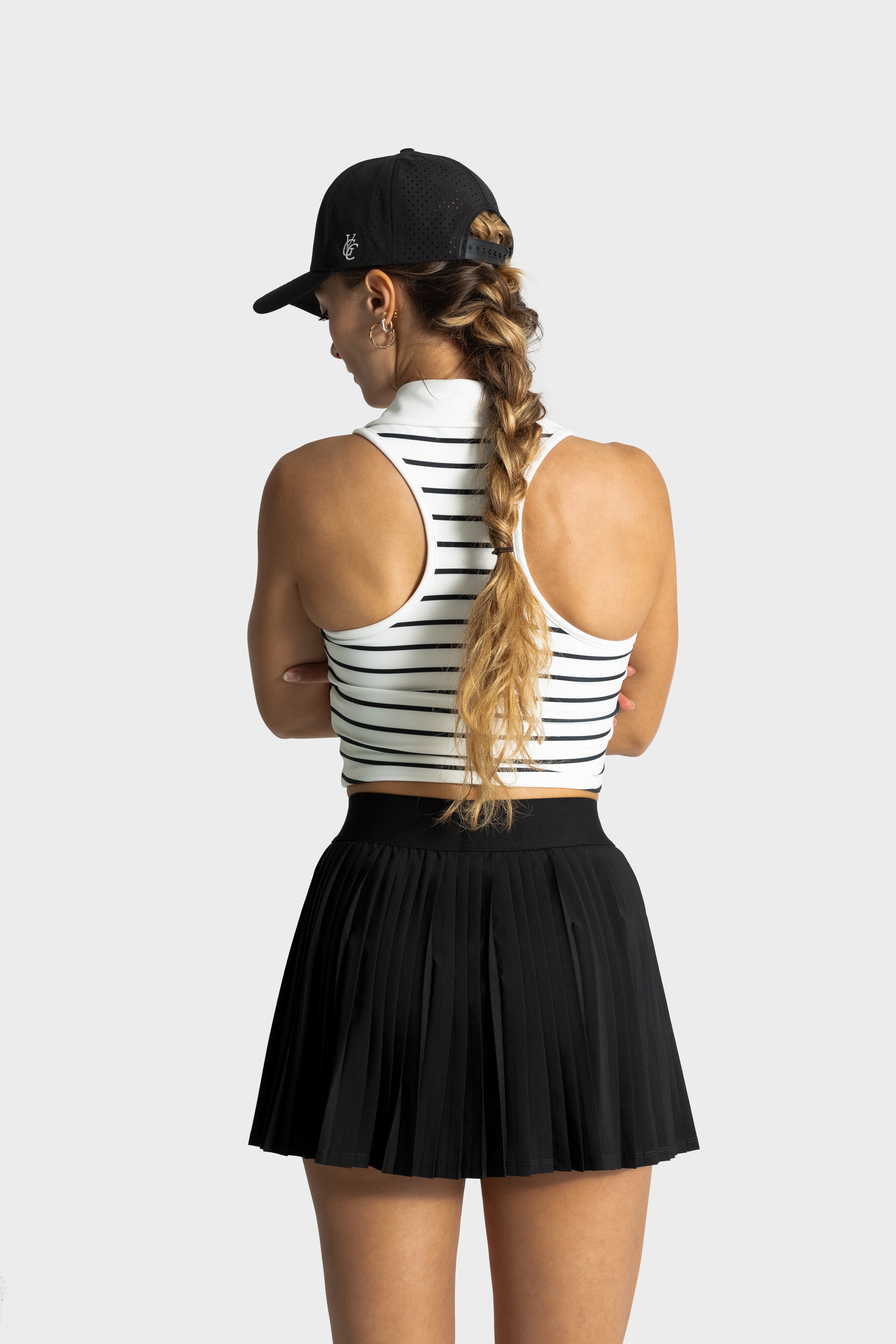 Woman wearing Perfetta golf skirt by Vittoria Golf Co with a black cap and striped tank top, standing against a gray background