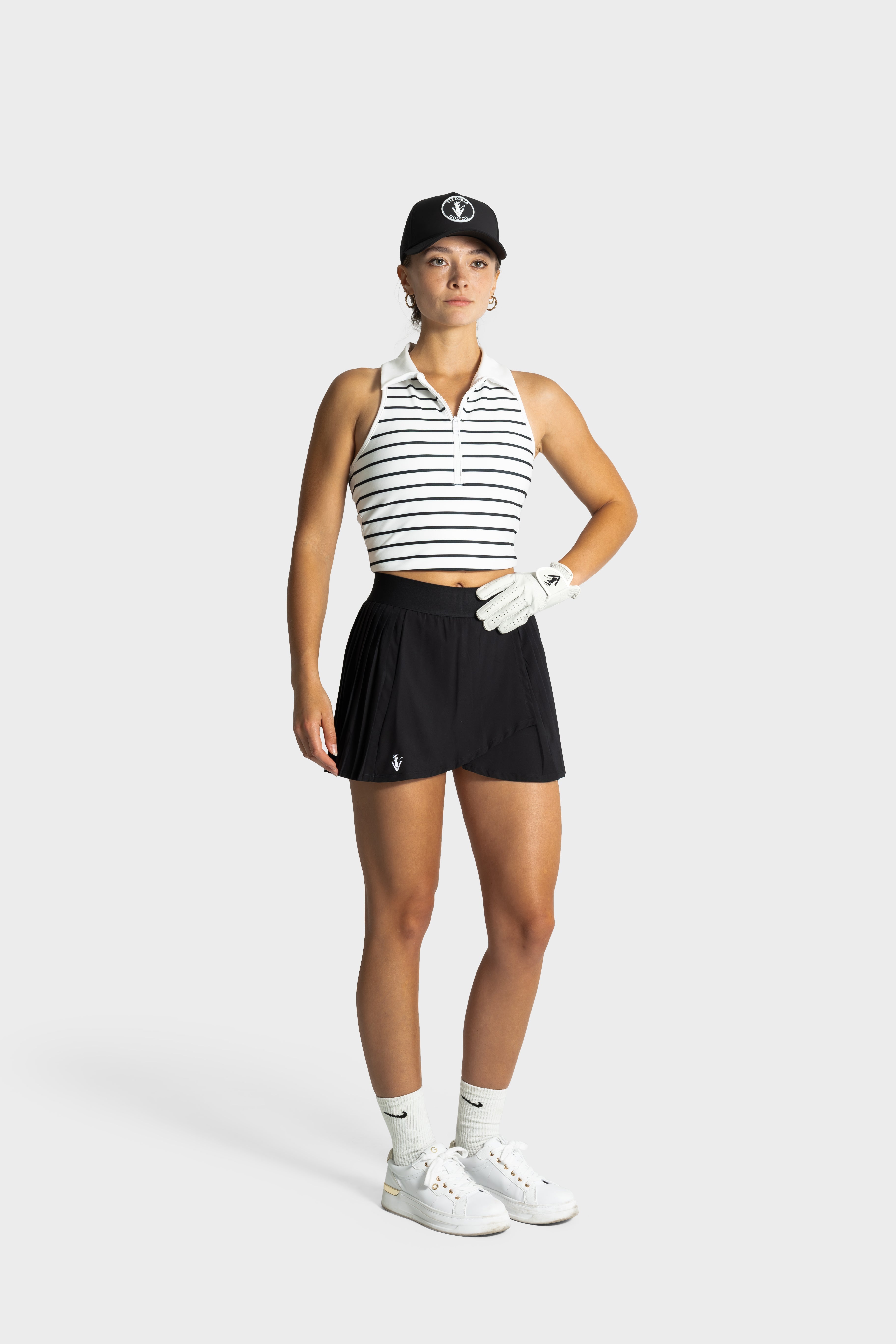 Woman wearing black Perfetta golf skirt with built-in shorts, white sleeveless striped polo, golf glove, and cap.