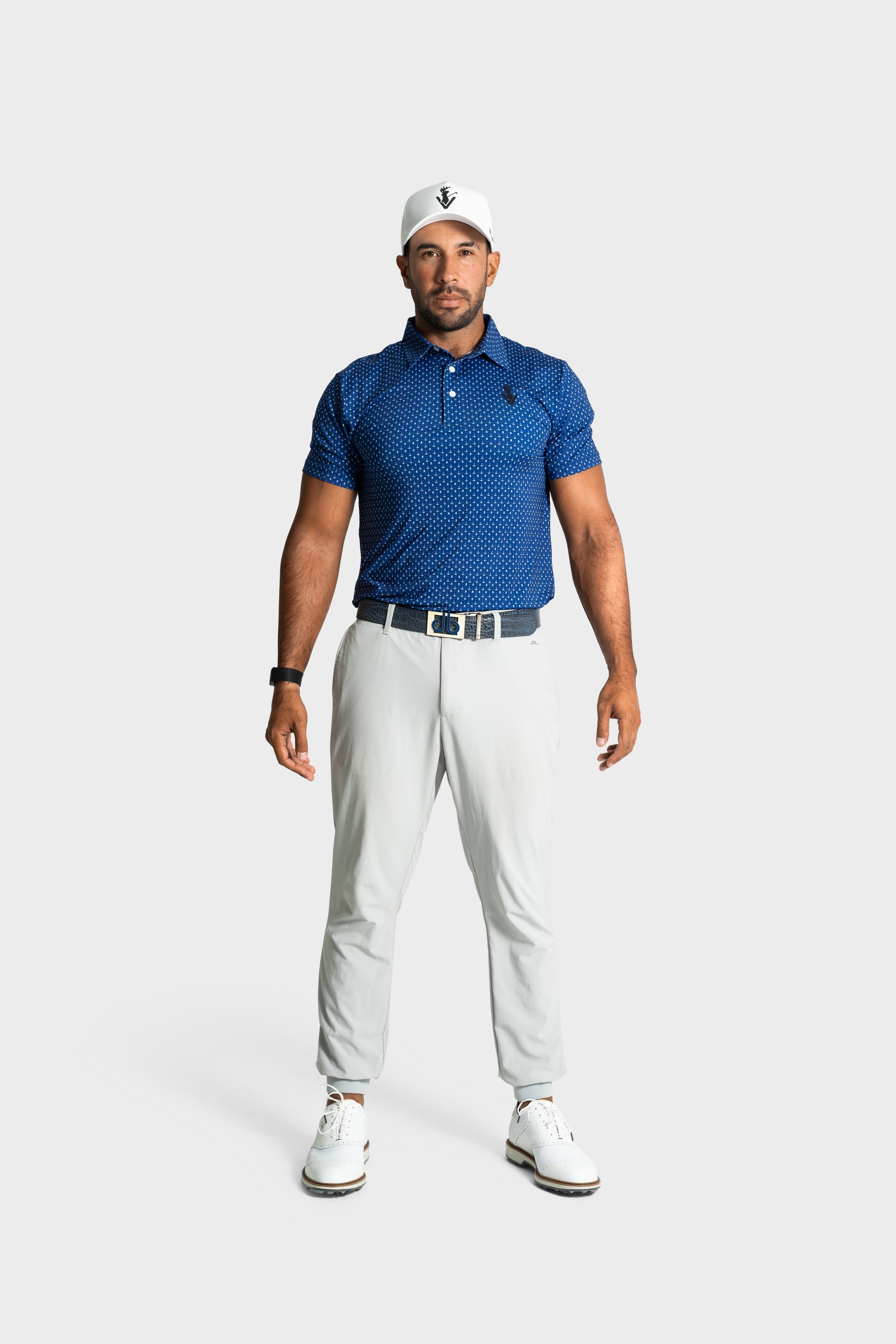 Male model wearing blue The Deadman polo with white golf pants and cap on white background