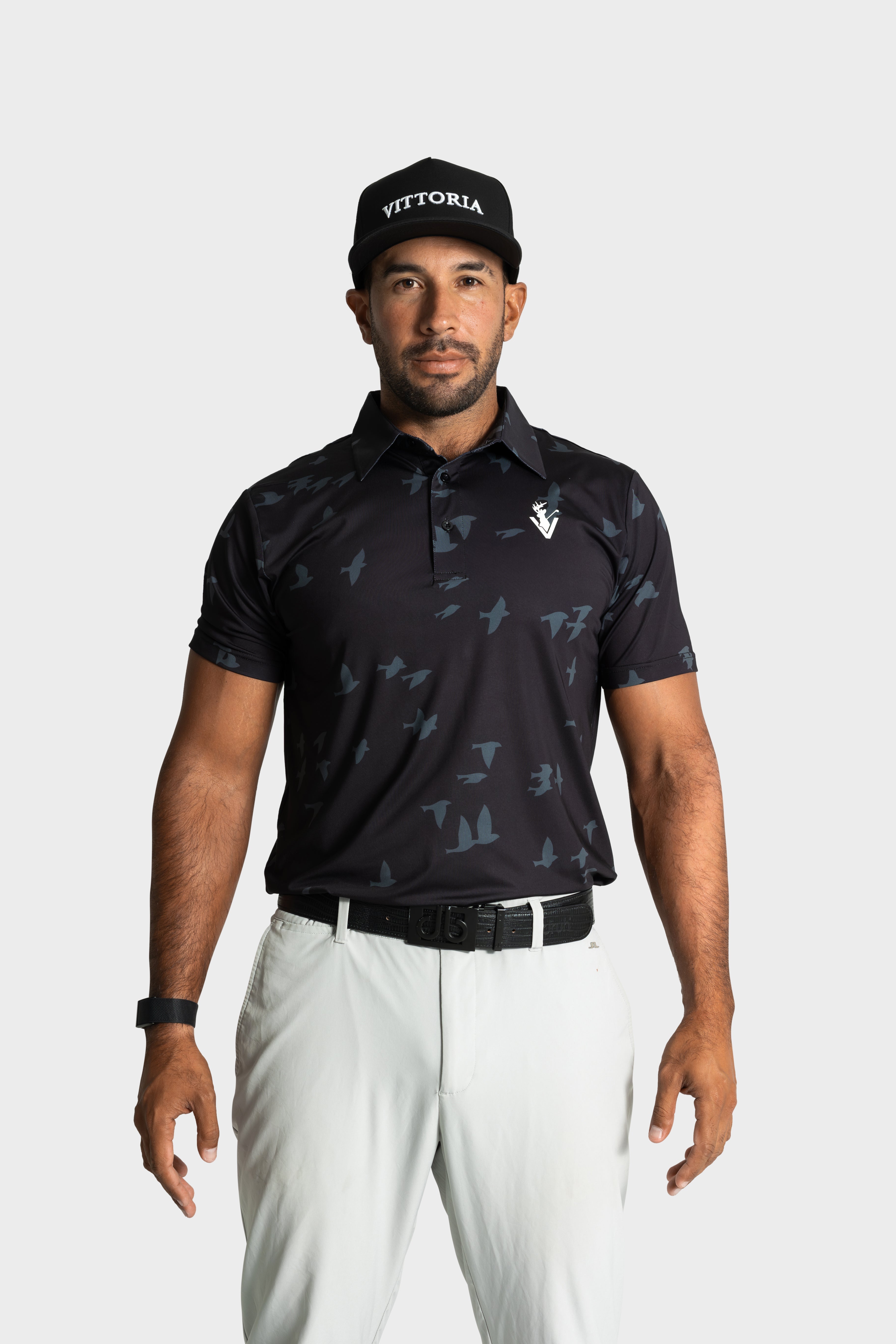 Male model wearing black The Flock polo with bird patterns and white pants by VittoriaGolfco