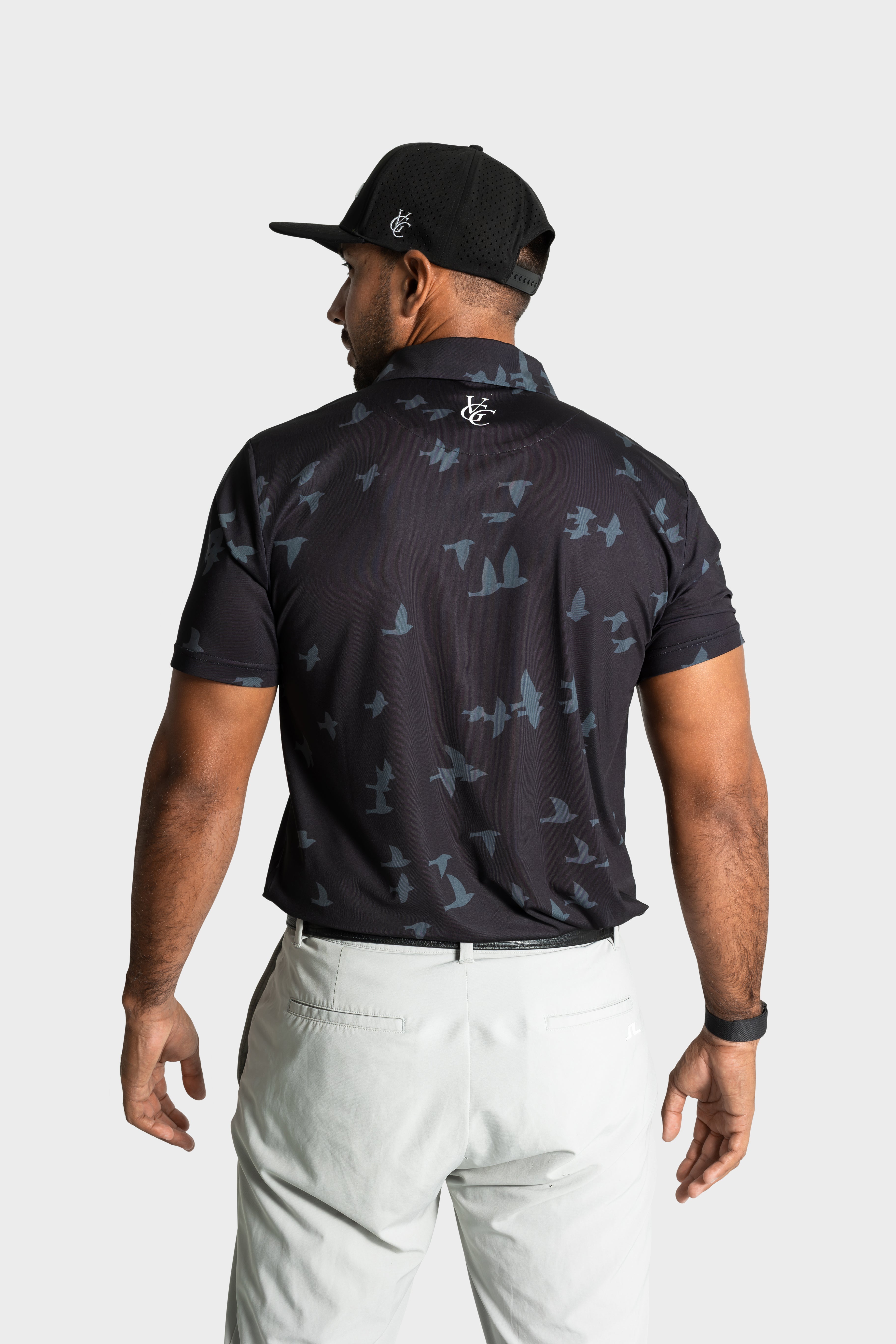 Man wearing black The Flock polo with bird pattern and white pants, back view