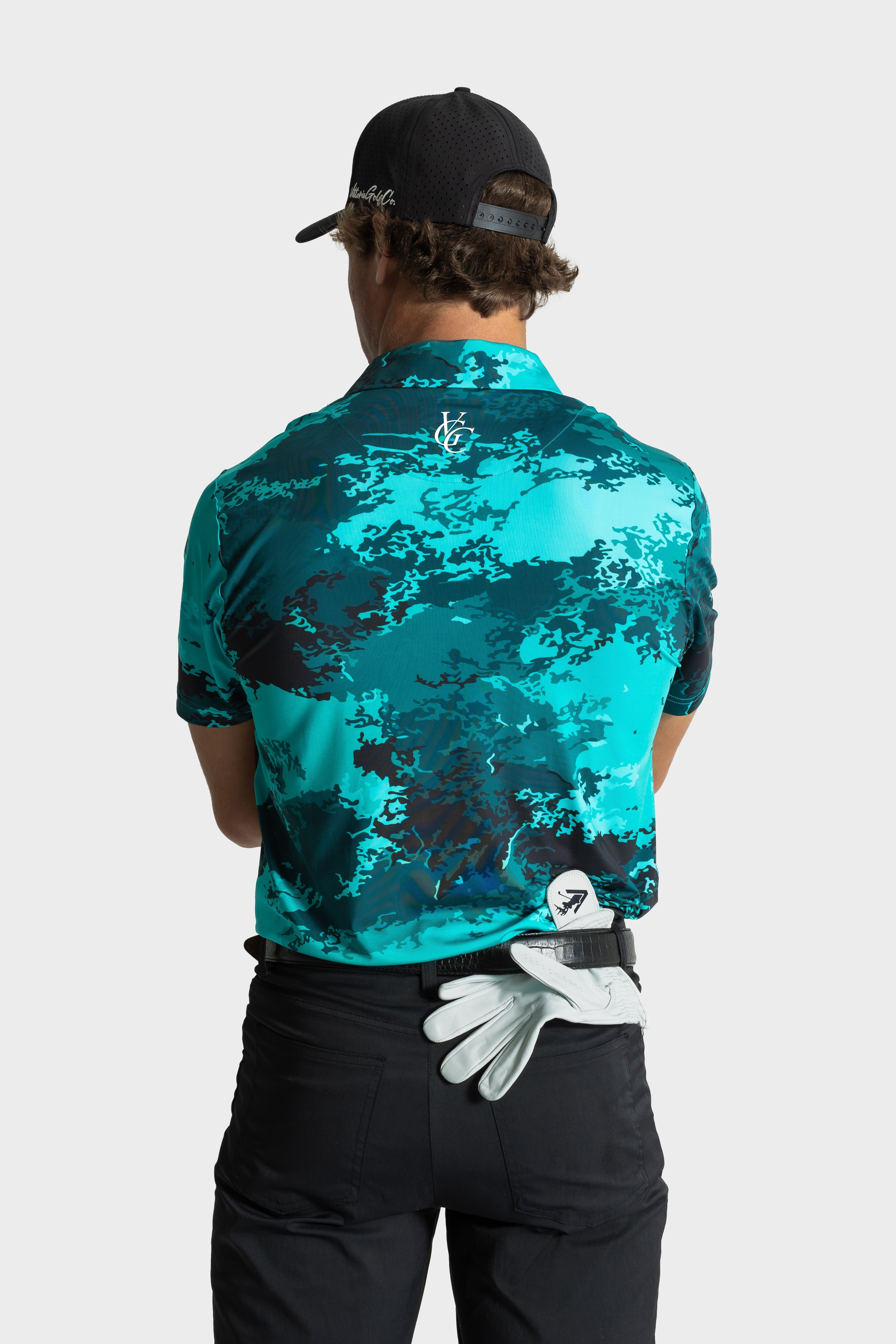 Back view of man wearing The One polo by VittoriaGolfco in teal camo pattern with black cap and golf glove tucked in pants