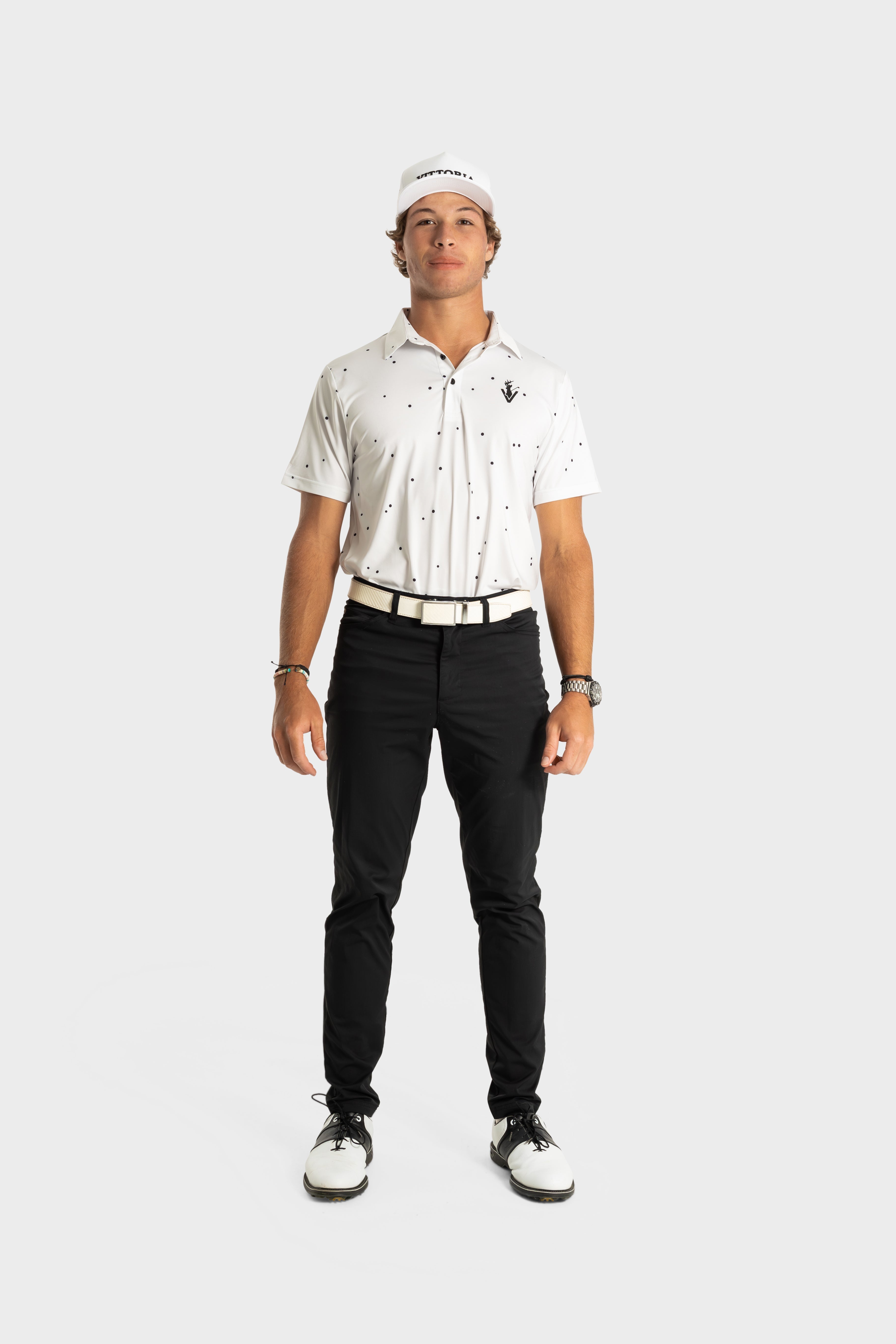 Male model wearing The Hinson polo white golf shirt with black pants and white golf shoes against a plain background