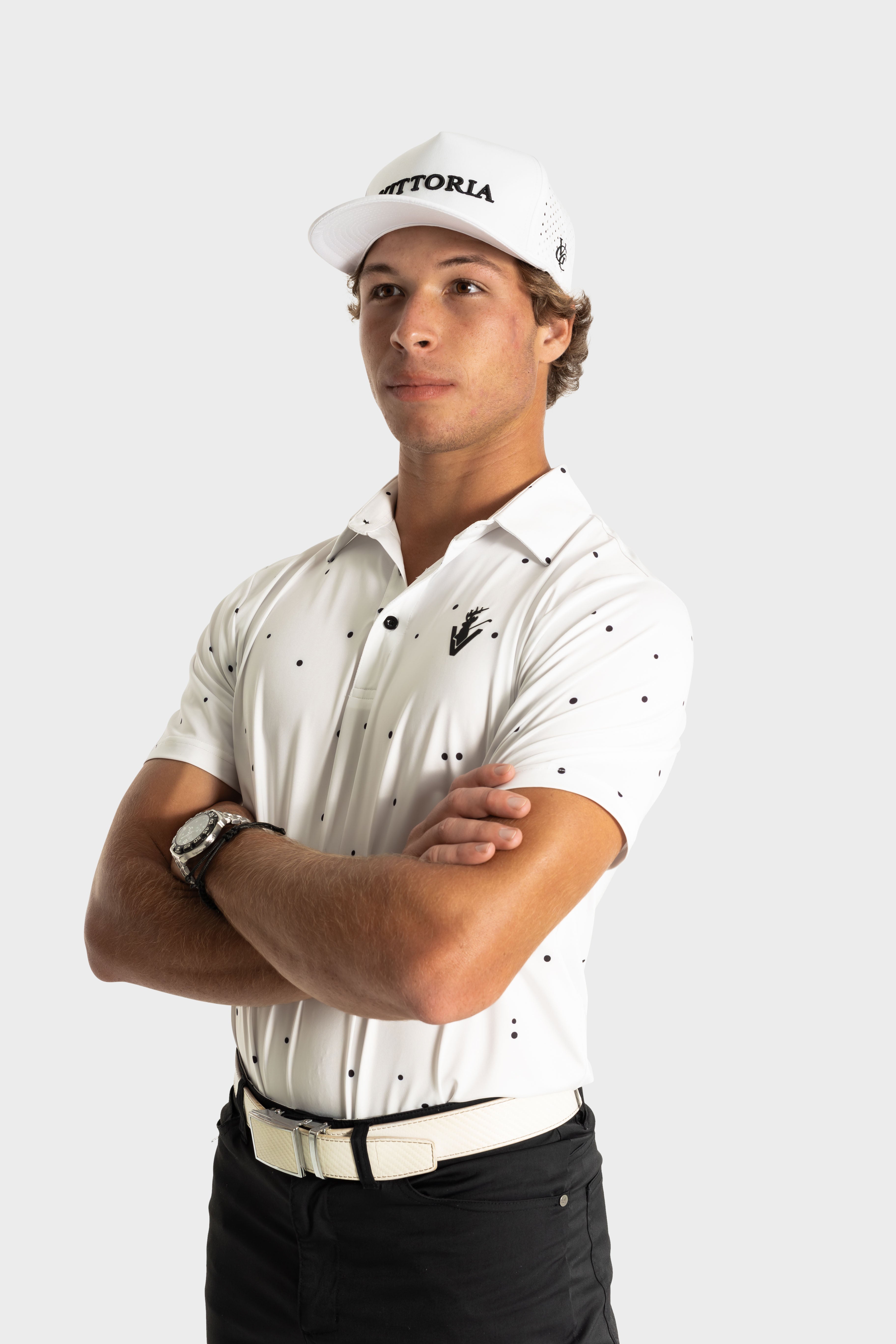 Man wearing The Hinson polo by VittoriaGolfco, white with small black dots, styled with a cap and black pants