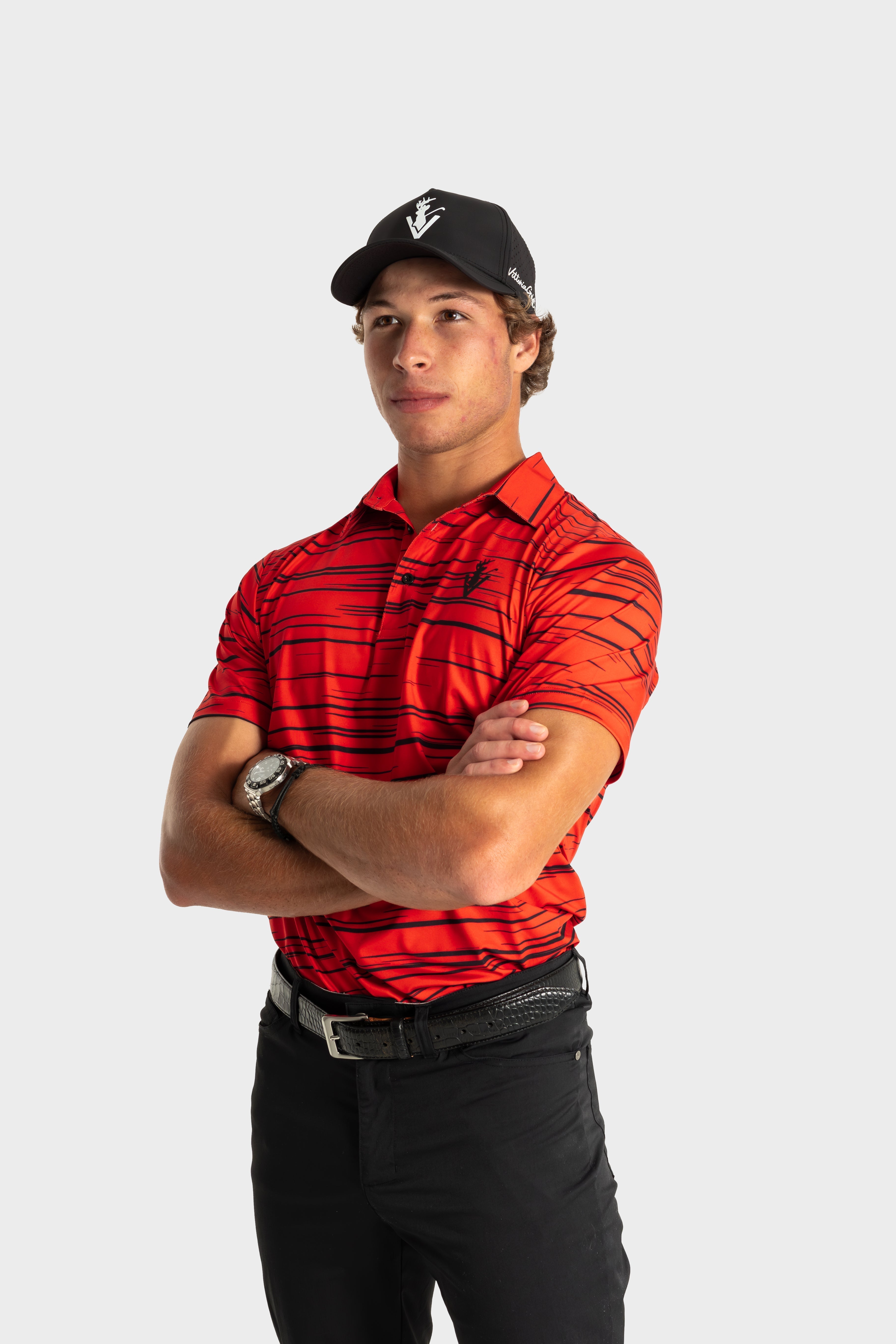 Man wearing red striped The Sunday Polo by VittoriaGolfCo with black pants and cap, standing confidently with arms crossed