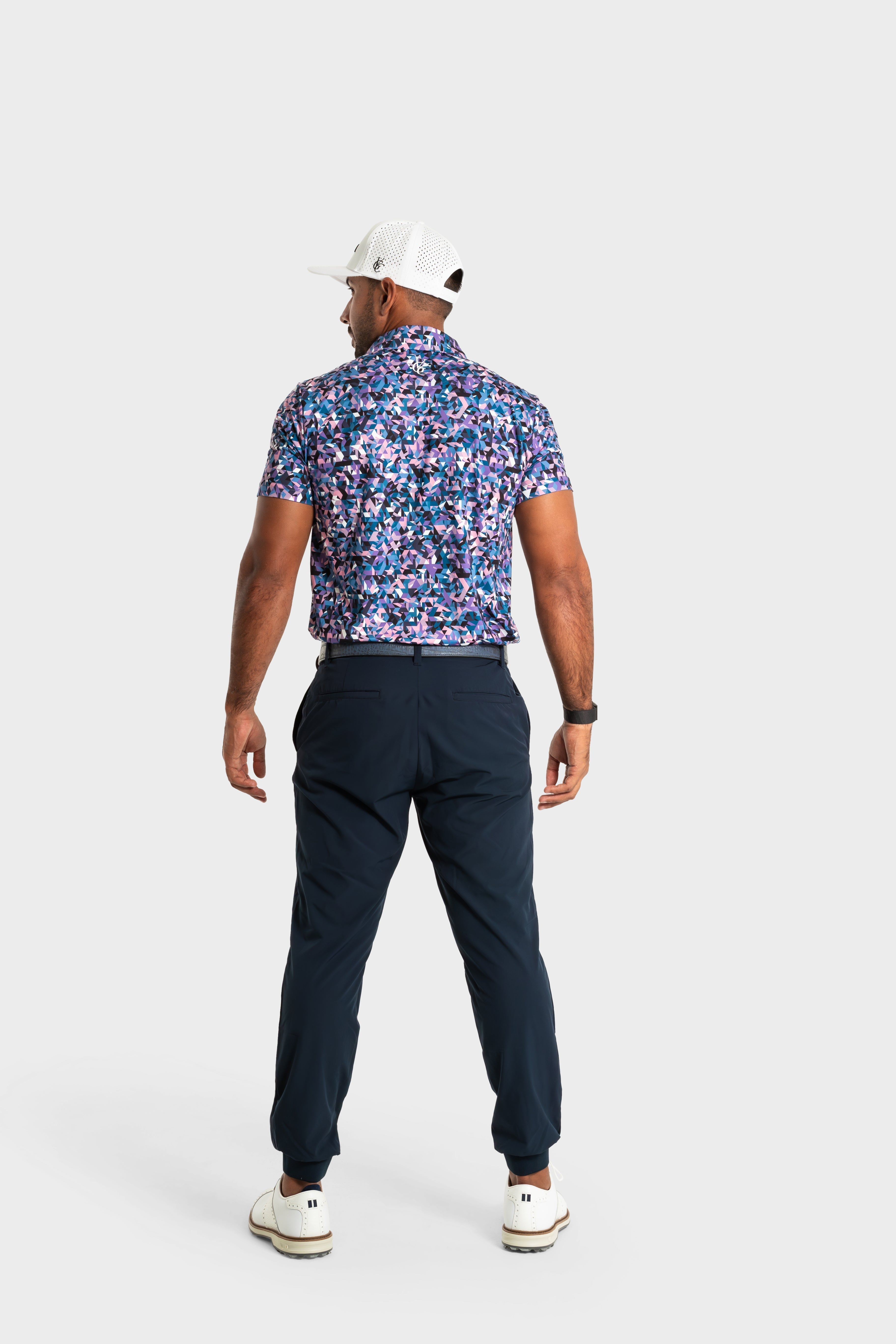 Man wearing The Geo polo with geometric patterns, navy pants, white cap, and sneakers, standing facing away