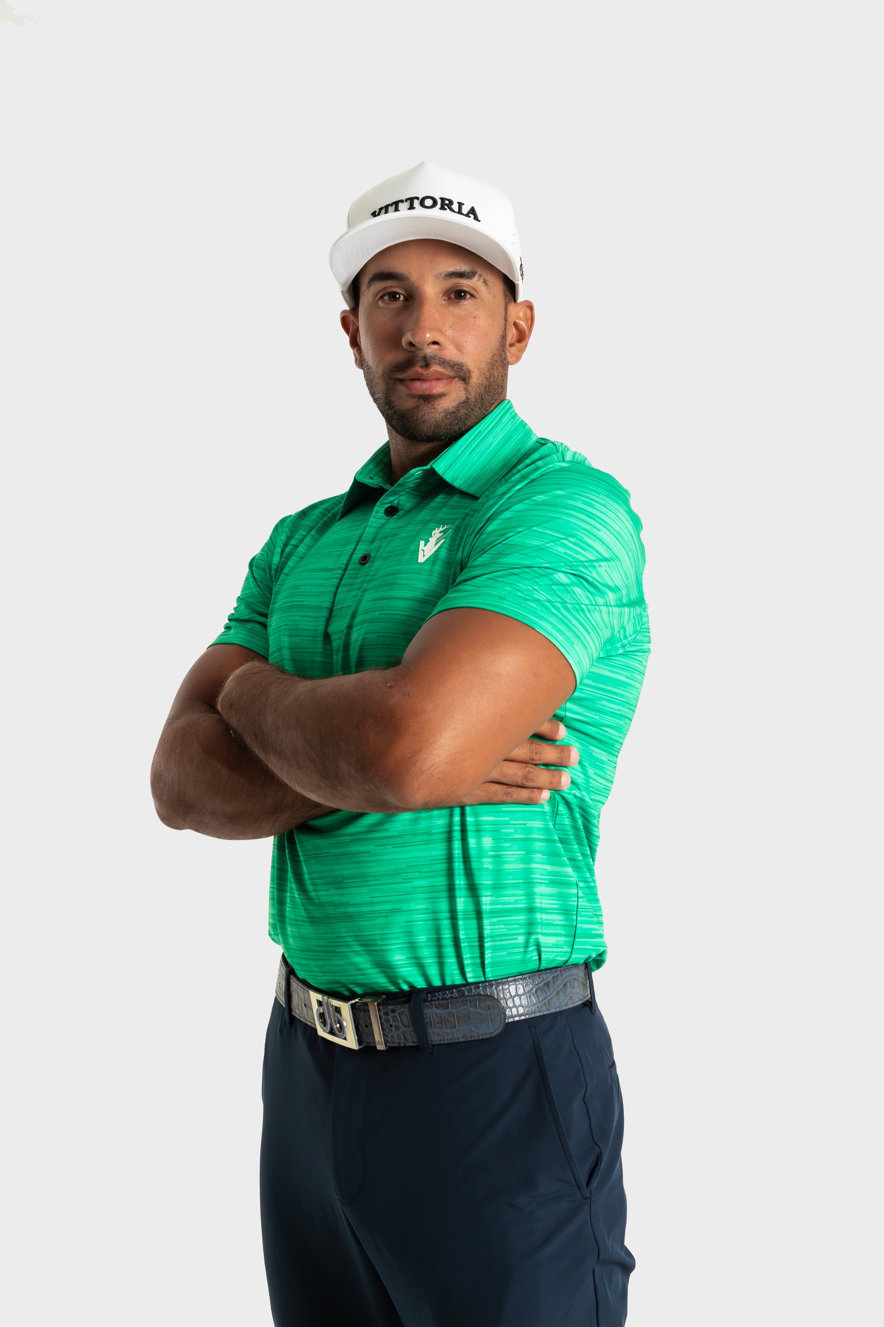 Man wearing green The Pasture polo by VittoriaGolfco with white cap and navy pants standing with arms crossed