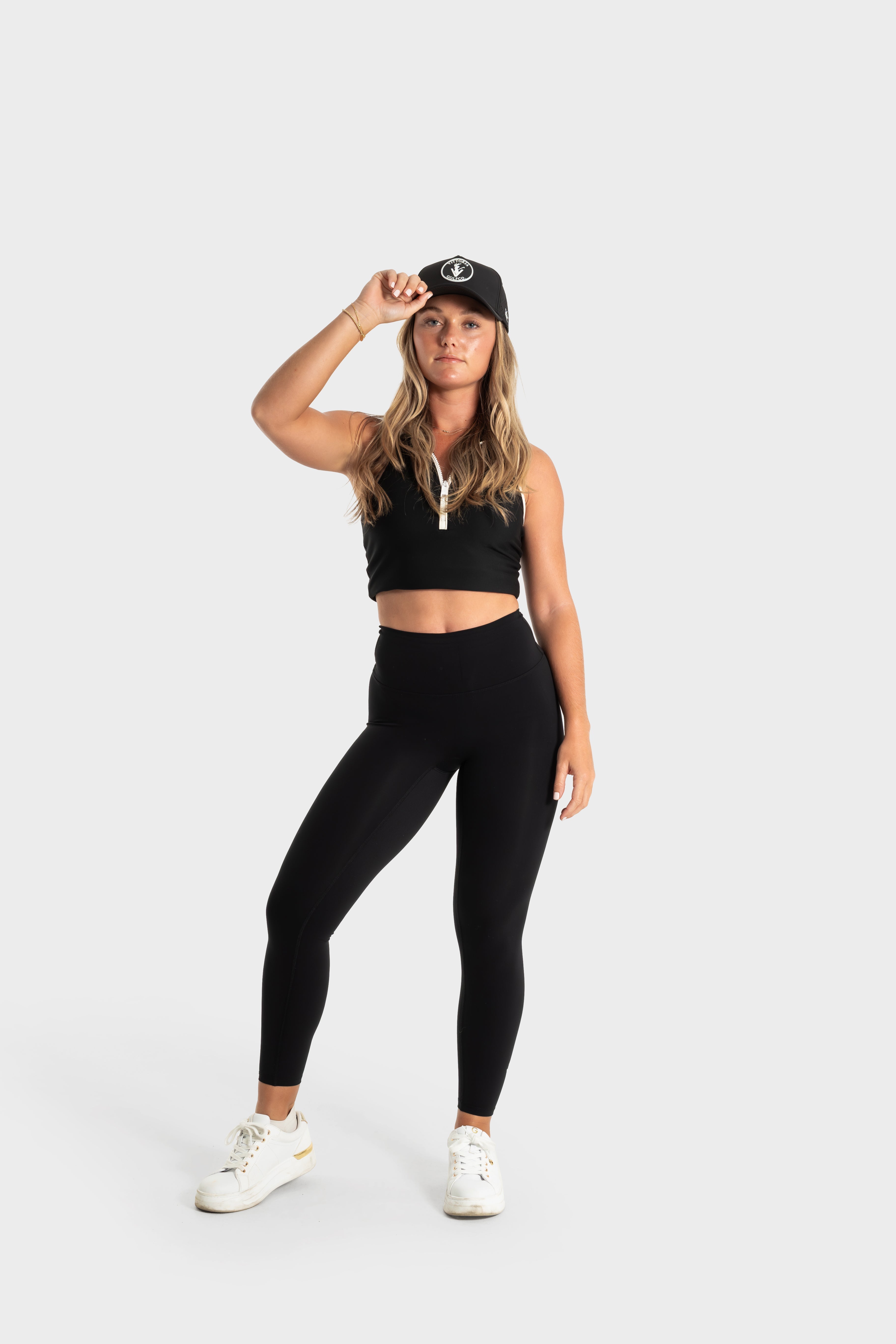 Woman wearing The Quelles black leggings and black crop top with cap, standing on white background
