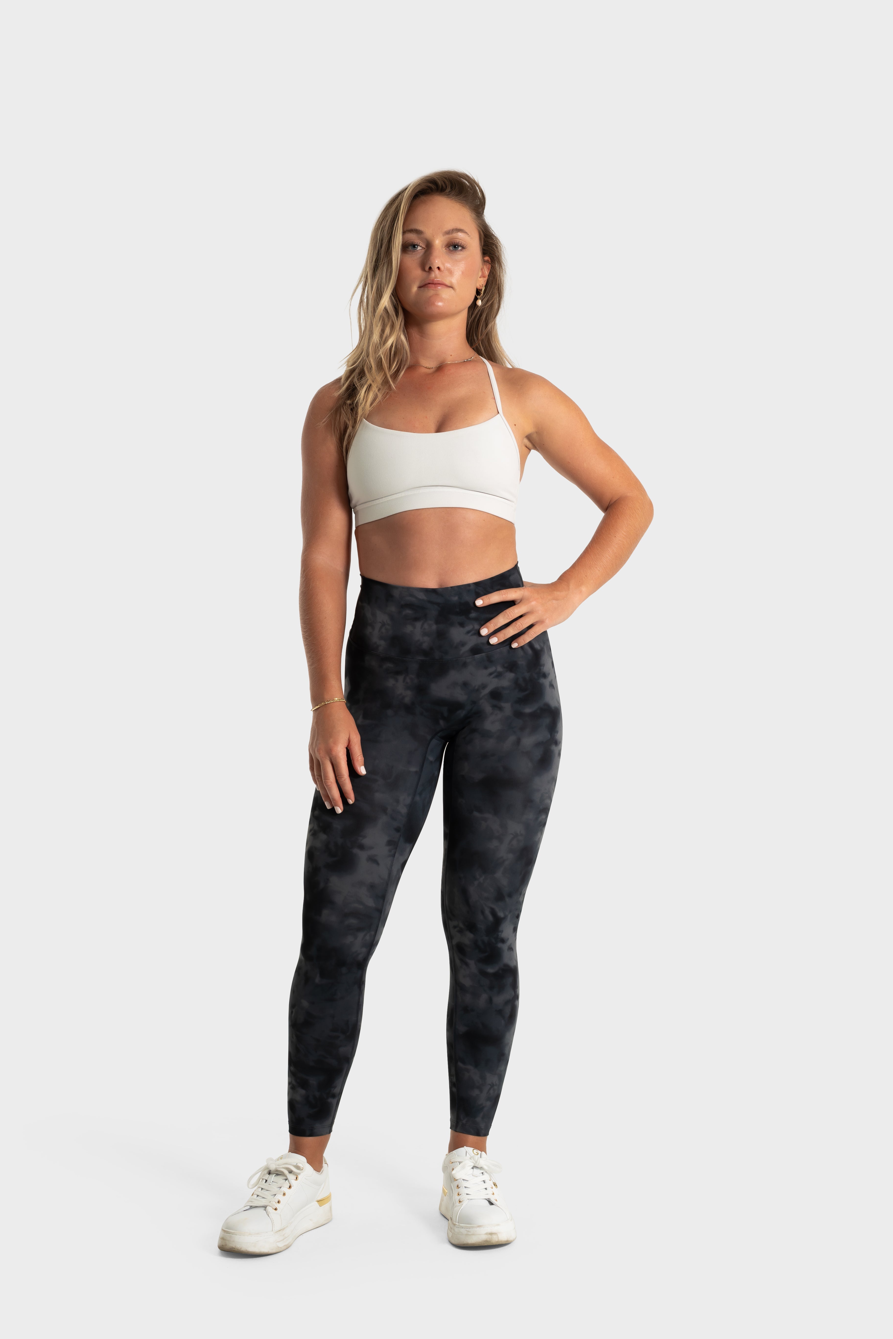 Woman wearing The Quelles black patterned leggings and white sports bra standing against a plain background