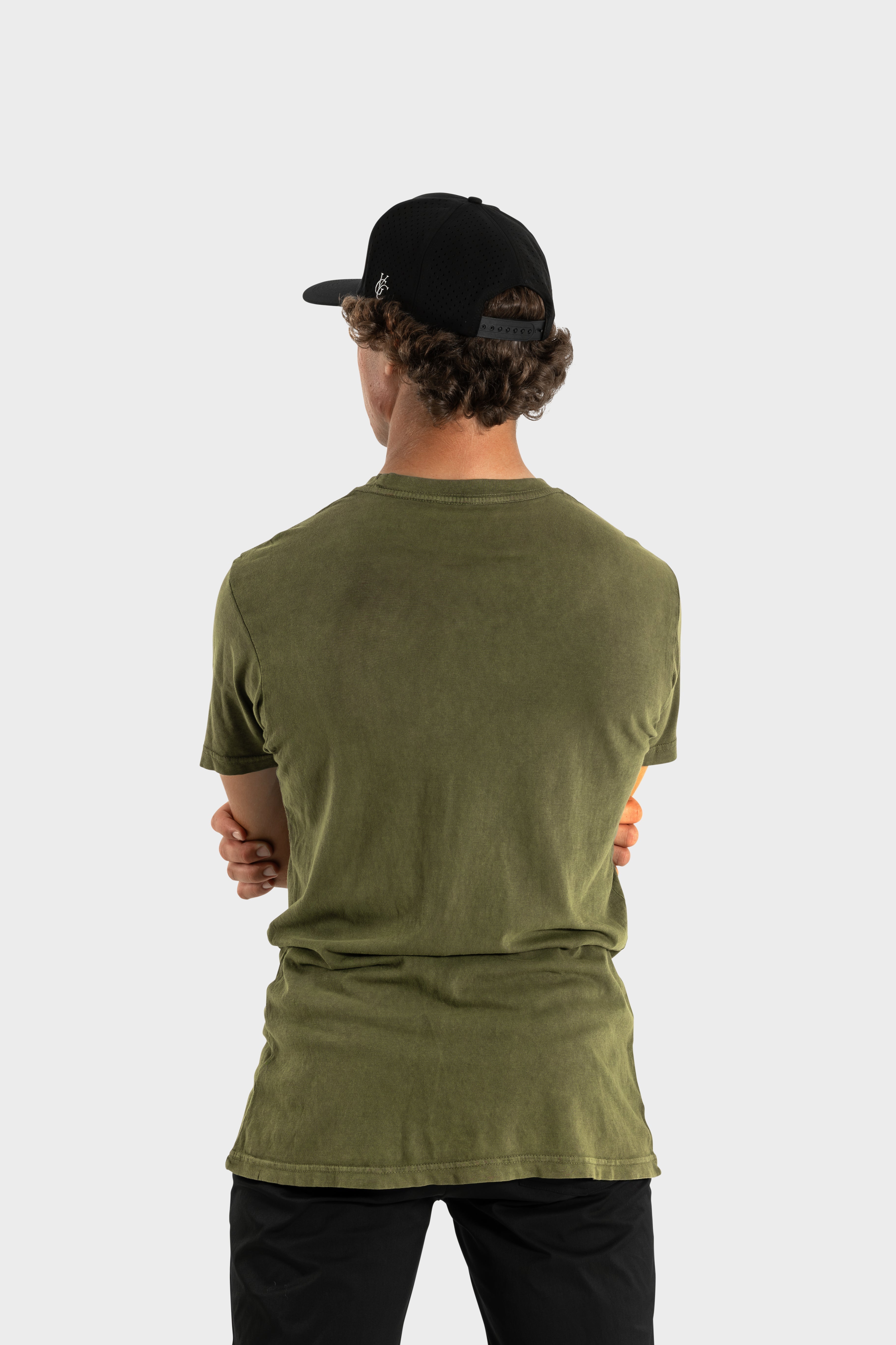 Back view of man wearing olive green Vittoria Oversized Tee with black cap and pants against white background