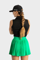 Woman wearing a green Perfetta golf skirt with pleats and a black sleeveless top and cap, showcasing the skirt's elegant and athletic design.