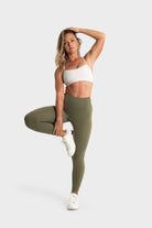 Active woman wearing The Quelles olive green leggings and white sports bra stretching against white background